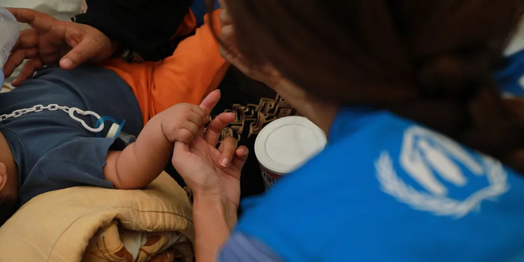 A UNHCR officer with her back to the camera holds the hand of an infant
